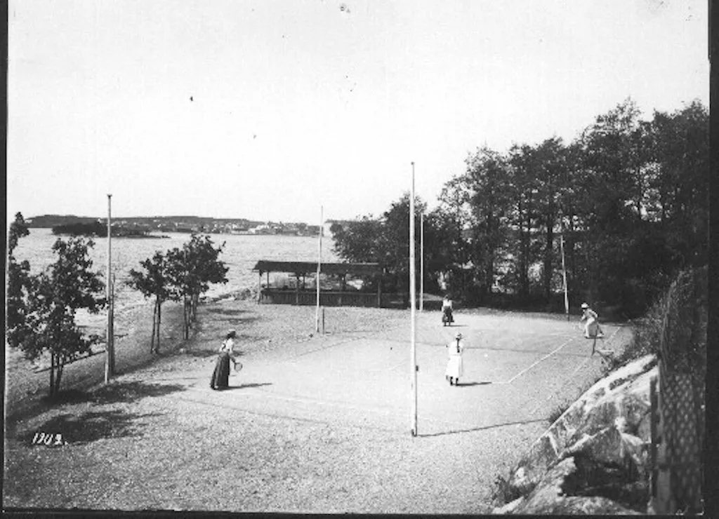 Gammalt fotografi av en grusbana vid vattnet med spelare i långa klänningar.